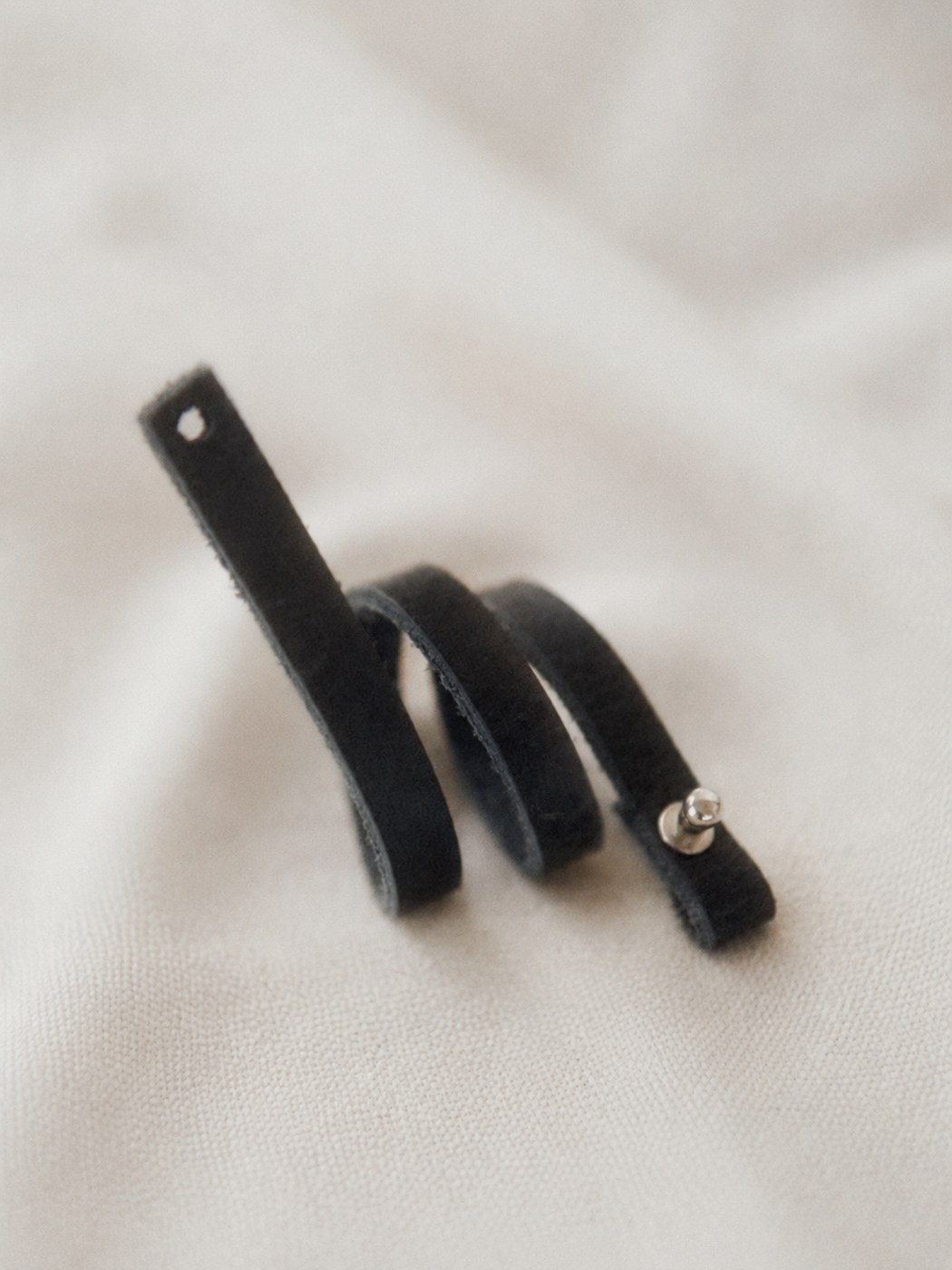 A blackened leather tie coiled on linen, closed with a chrome post.
